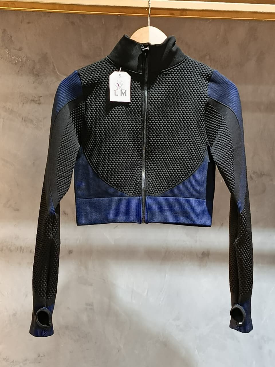 Chaqueta corta negra y azul con cremallera, colgada de una barra de madera.