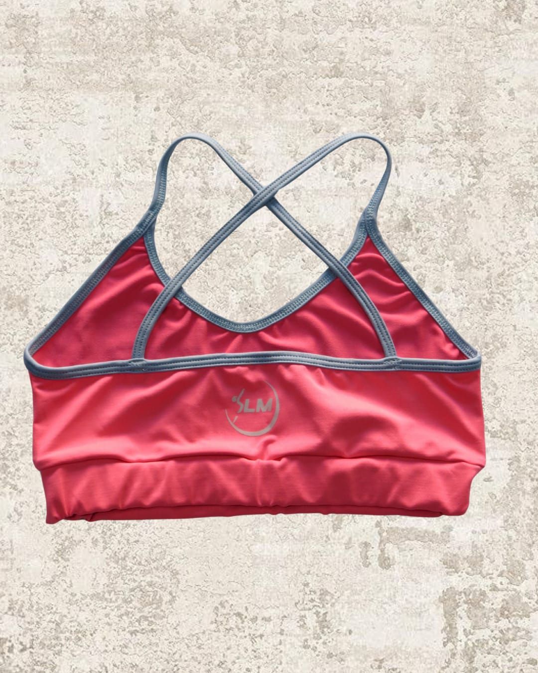 Sujetador deportivo rosa con ribete gris y tirantes cruzados, sobre un fondo texturizado.