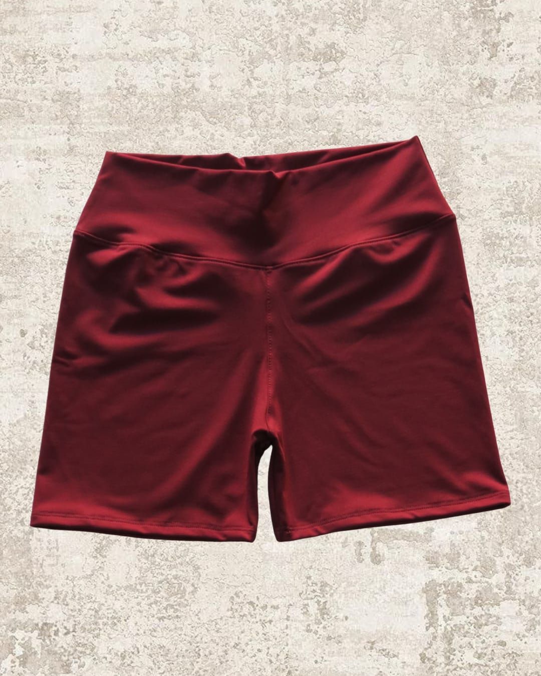 Pantalones cortos deportivos de cintura alta de color rojo oscuro sobre un fondo marrón claro texturizado.