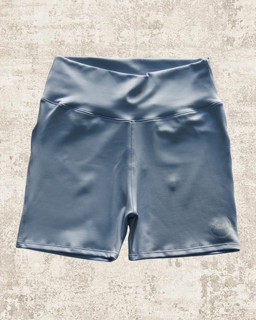 Pantalón corto deportivo de color azul grisáceo con cintura alta, sobre una superficie texturizada de color claro.
