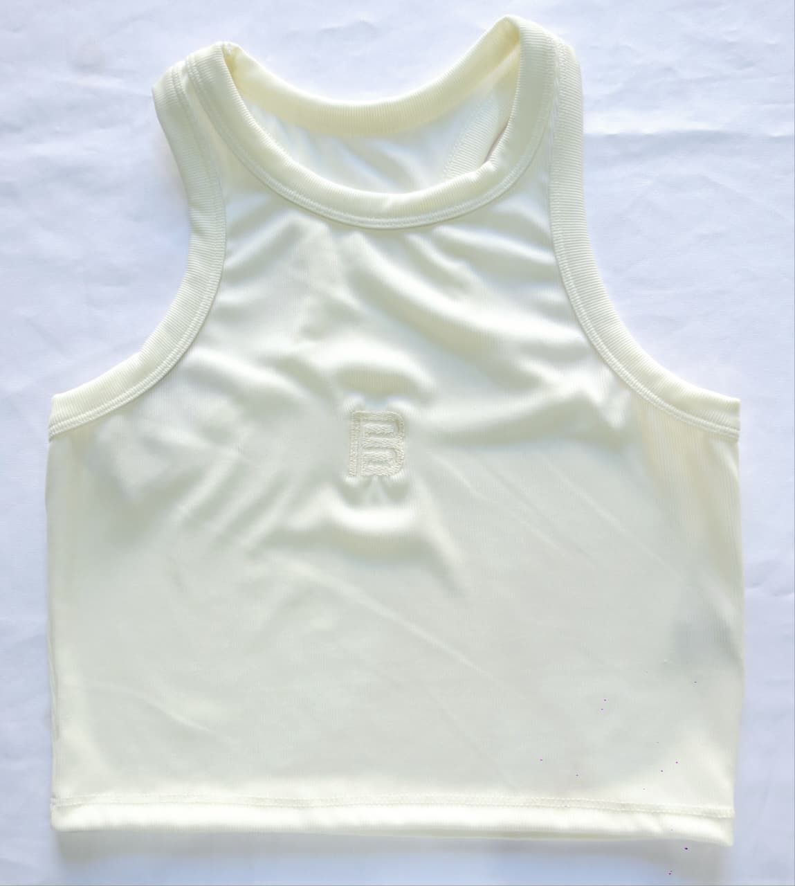 Camiseta interior sin mangas de color crema con cuello acanalado y un pequeño diseño en relieve cerca del centro.