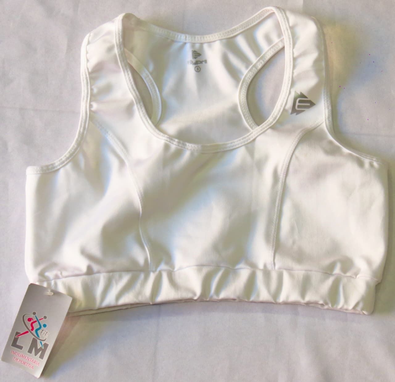 Sujetador deportivo blanco con espalda nadadora y logotipo, doblado sobre una superficie con etiqueta.