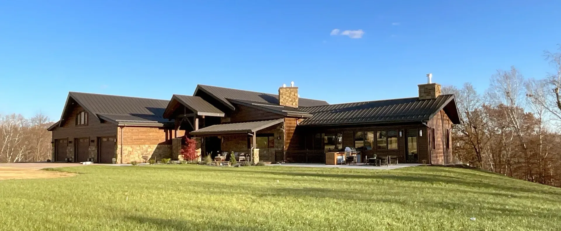 A large rustic-style house with a dark roof and brick chimney on a grassy hill under a blue sky.