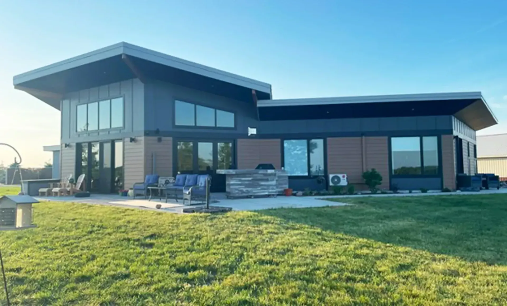 Modern house with dark gray and brown siding, large windows, and patio on a grassy lawn.