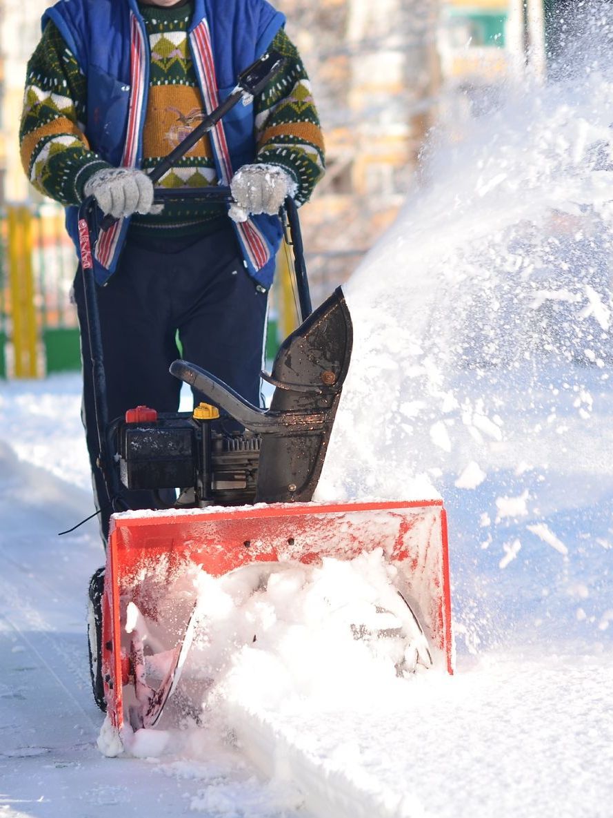 a man is blowing snow from a snow blower on a sidewalk .