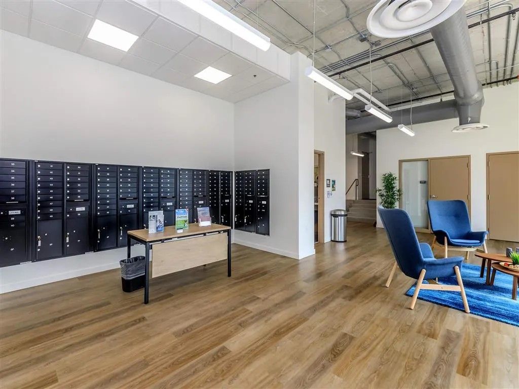 Lobby with mailboxes, seating area, and wooden floors. Blue armchairs, a table, and a stainless steel trash can are visible.