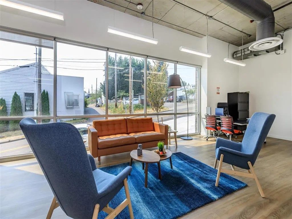 Modern office lounge: blue chairs, orange sofa, blue rug, large windows, and outdoor view.