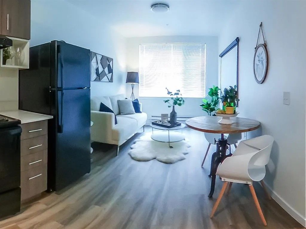 Small apartment interior with kitchen, living room, and dining area. Includes black refrigerator, white couch, round table.