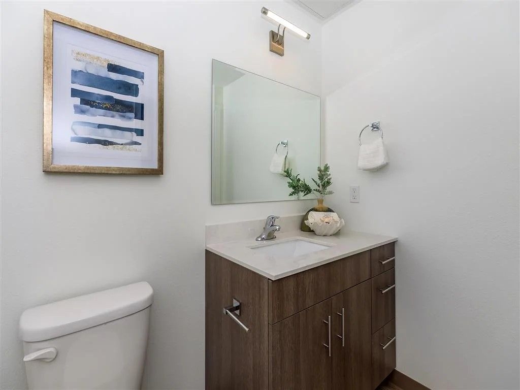 Small bathroom with toilet, sink, mirror, and artwork on a white wall.