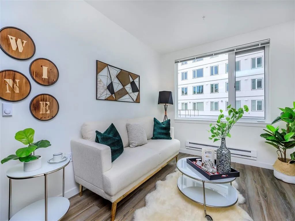 Living room with white sofa, artwork, plants, and window overlooking a building.