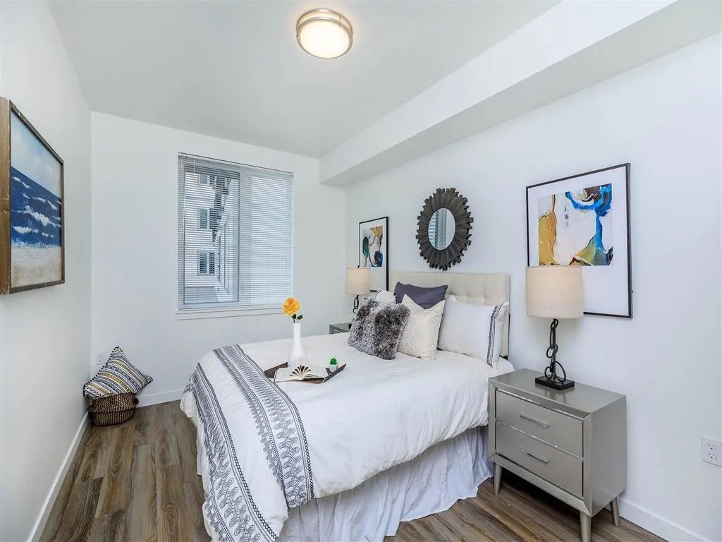Bedroom with white walls, window with blinds, bed with decorative pillows, nightstand.
