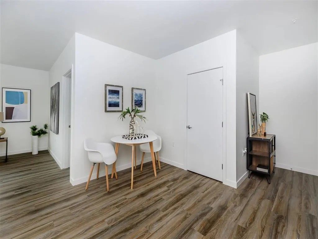 Brightly lit apartment interior with a small dining table, white chairs, and artwork. Wood flooring.