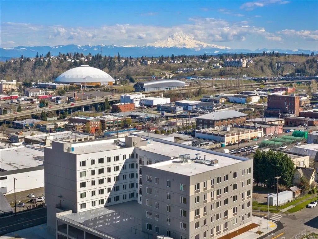 Cityscape with buildings, dome stadium, and mountain range under a bright blue sky.