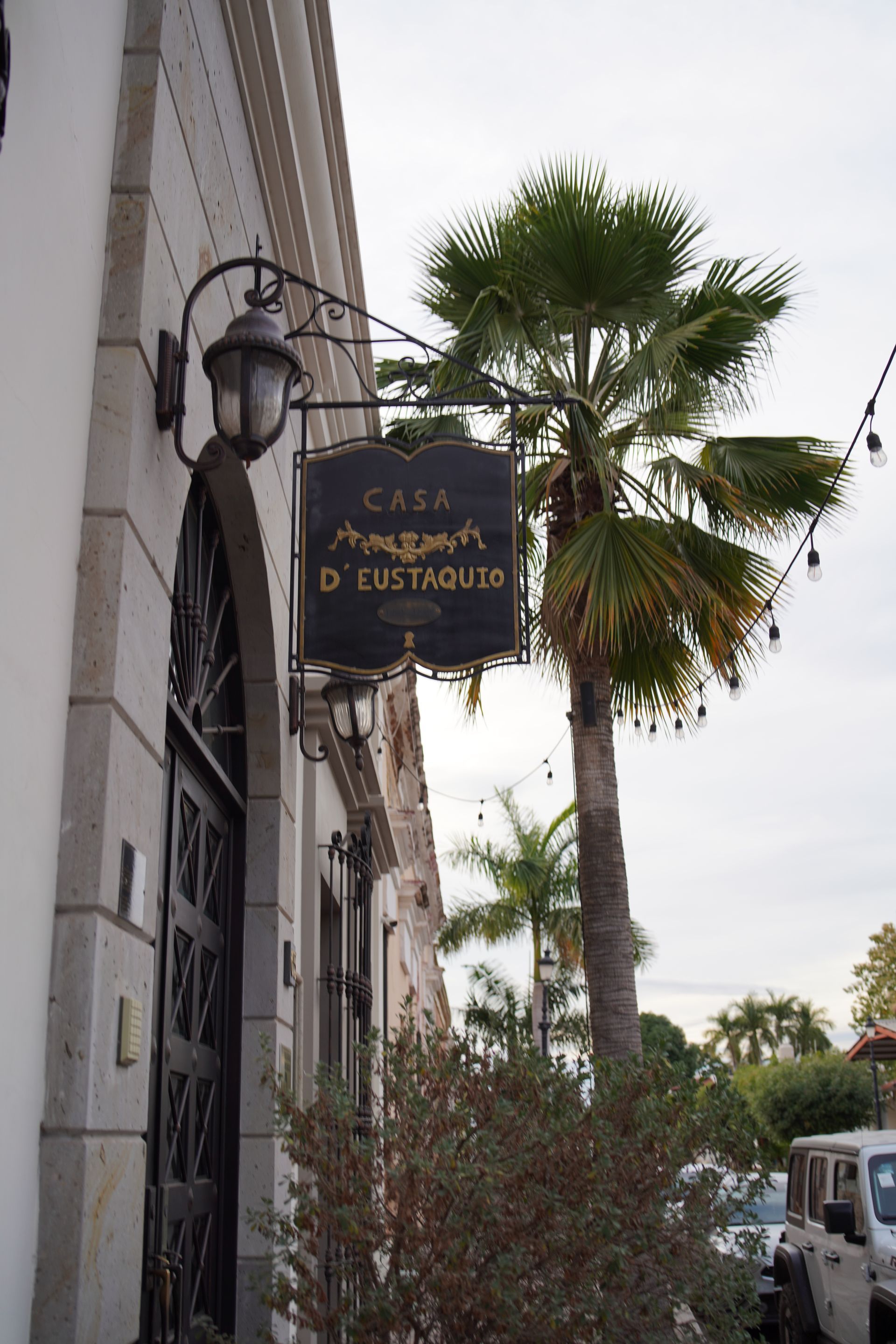 A building with a sign hanging from the side of it and a palm tree in front of it.