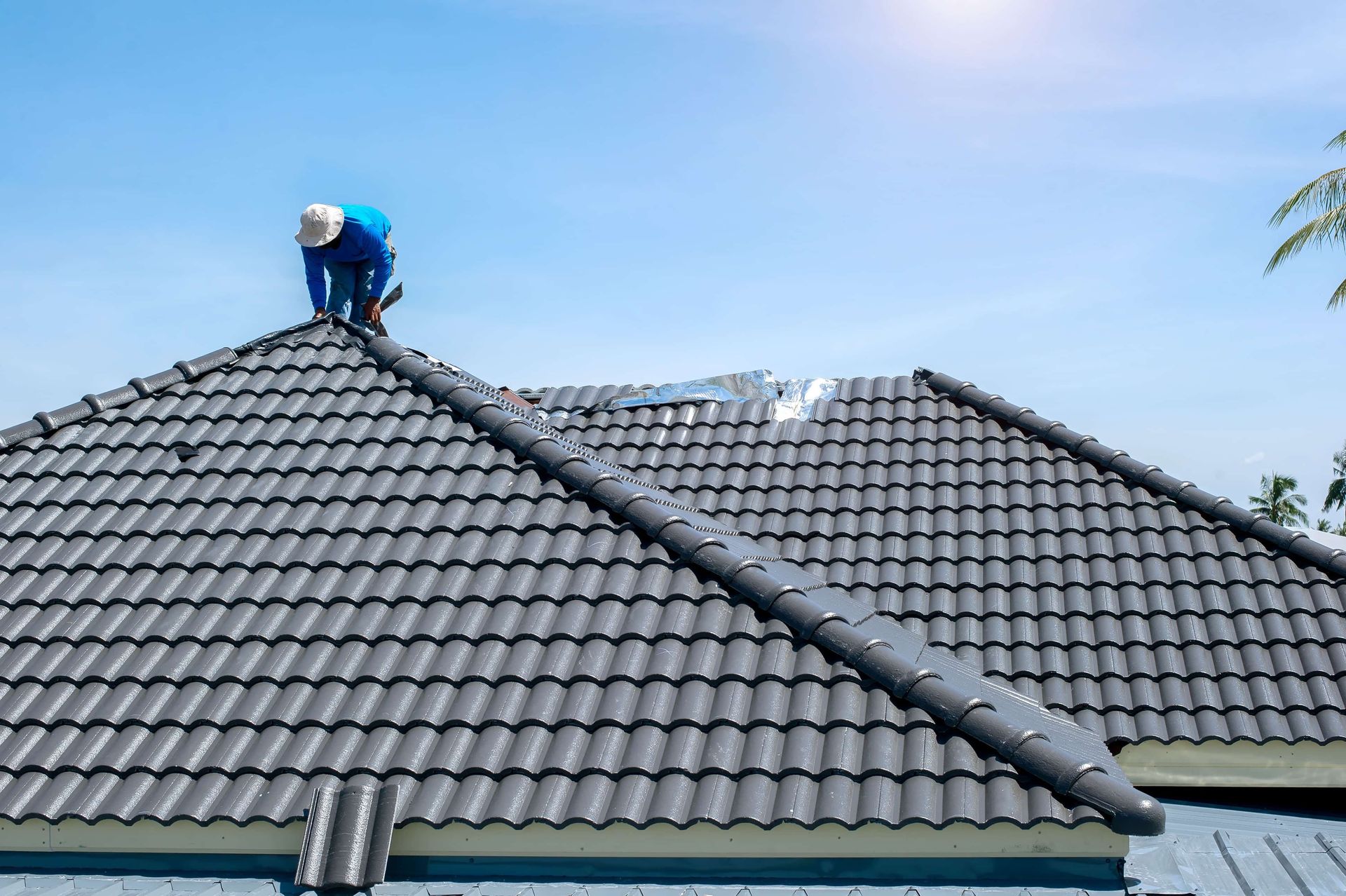 Worker on a gray tiled roof inspecting the ridge under a clear blue sky.
