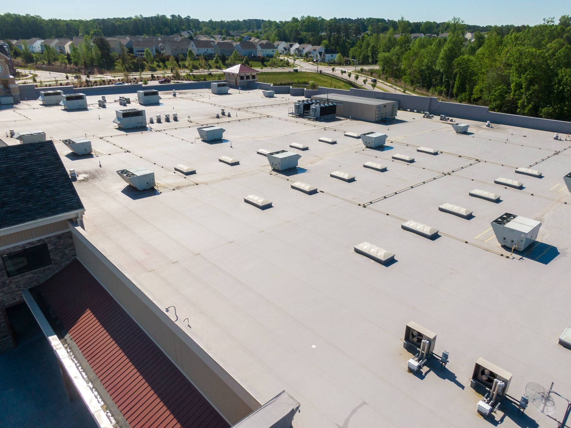 Aerial view of a large white flat rooftop with vents and equipment, bordered by trees and buildings.