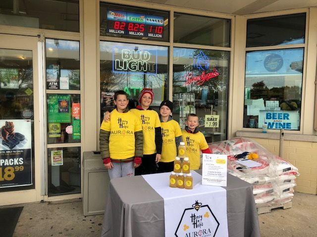 Four people in yellow shirts stand outside a liquor store beside stacked boxes and a table with merchandise.