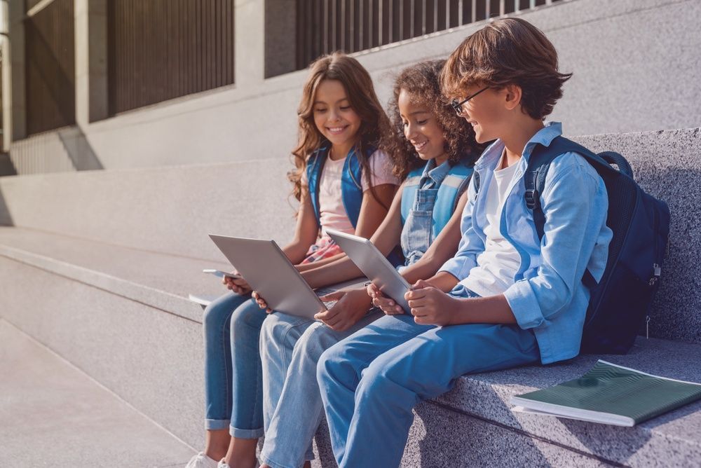 Three students sit on steps, smiling and working on a laptop outdoors.