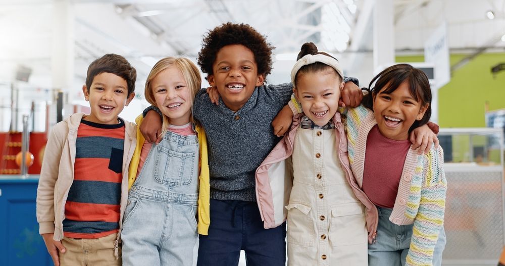 Five smiling children standing arm in arm in a bright indoor hallway