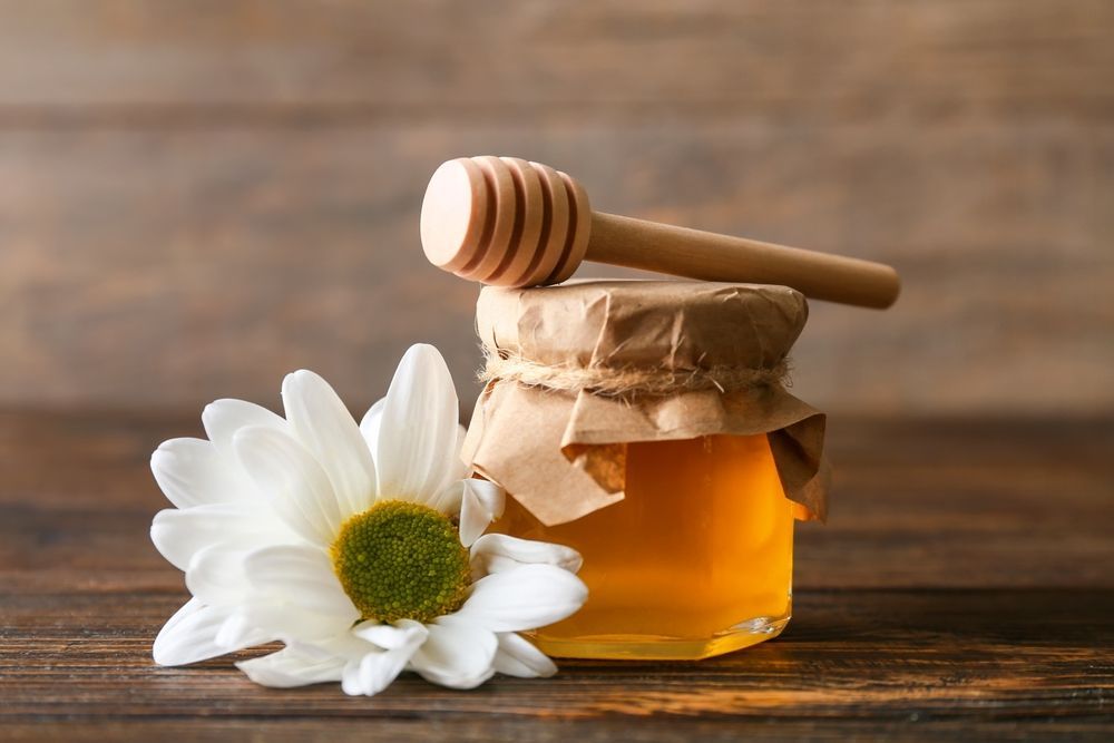 Jar of honey with a honey dipper on top, a flower to the side, on a wooden surface.
