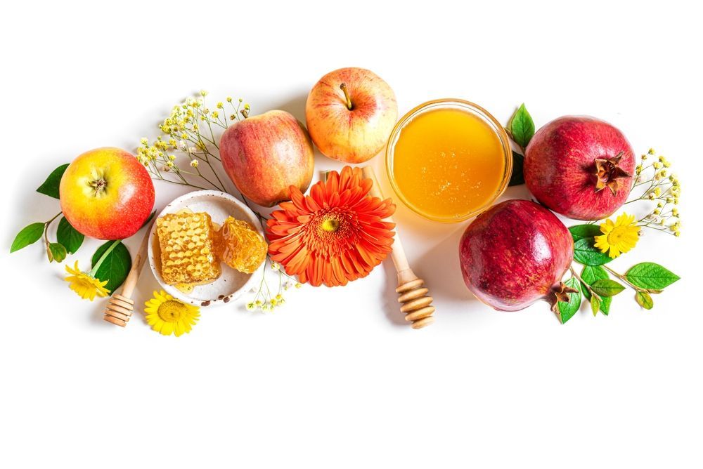 Apples, pomegranates, honey, and honeycomb arranged on a white background, with flowers and wooden honey dippers.