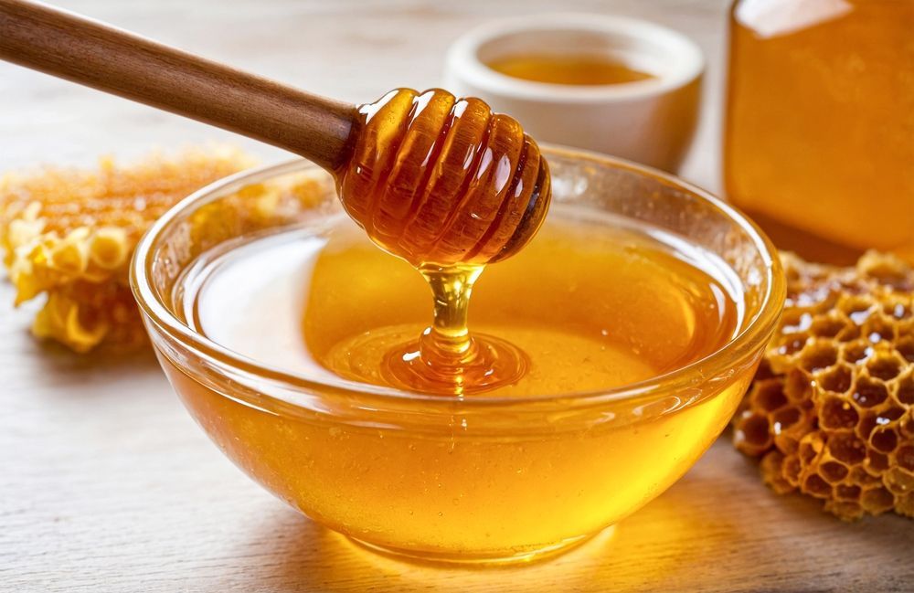 Honey dripping from a wooden dipper into a glass bowl, with honeycomb in the background.
