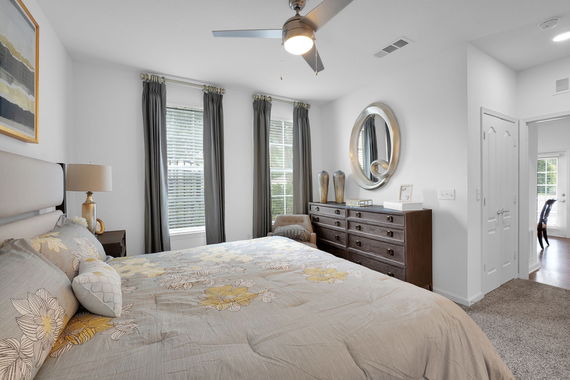 Bedroom with white walls, gray curtains, dark wood dresser, bed with gold accents.