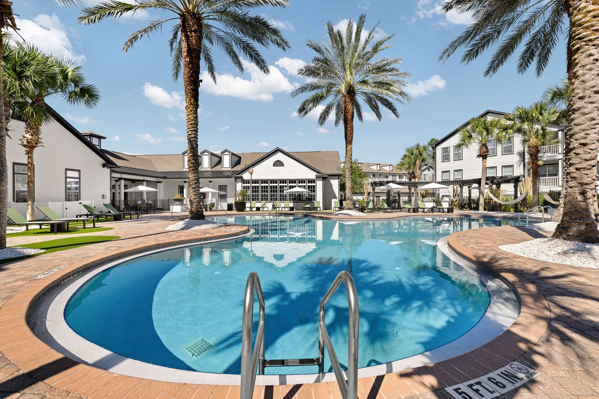 Swimming pool with steps, surrounded by brick patio, palm trees and buildings, on a sunny day.