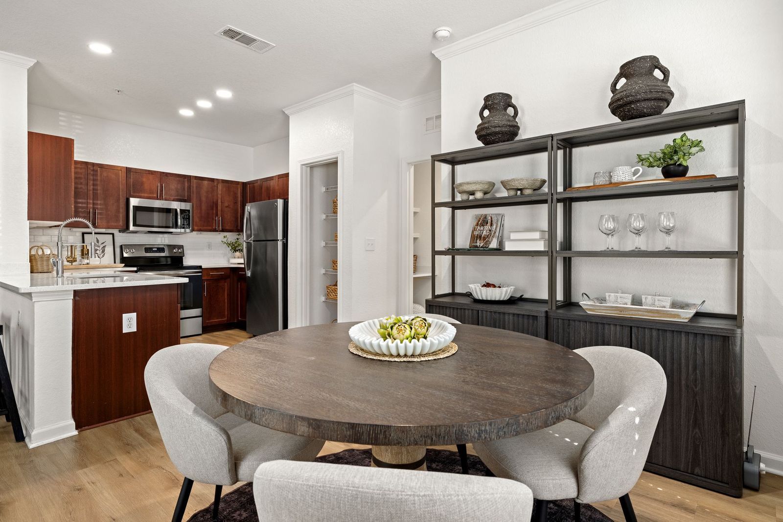 A round wooden dining table with four grey upholstered chairs, adjacent to an open kitchen with dark wood cabinetry.
