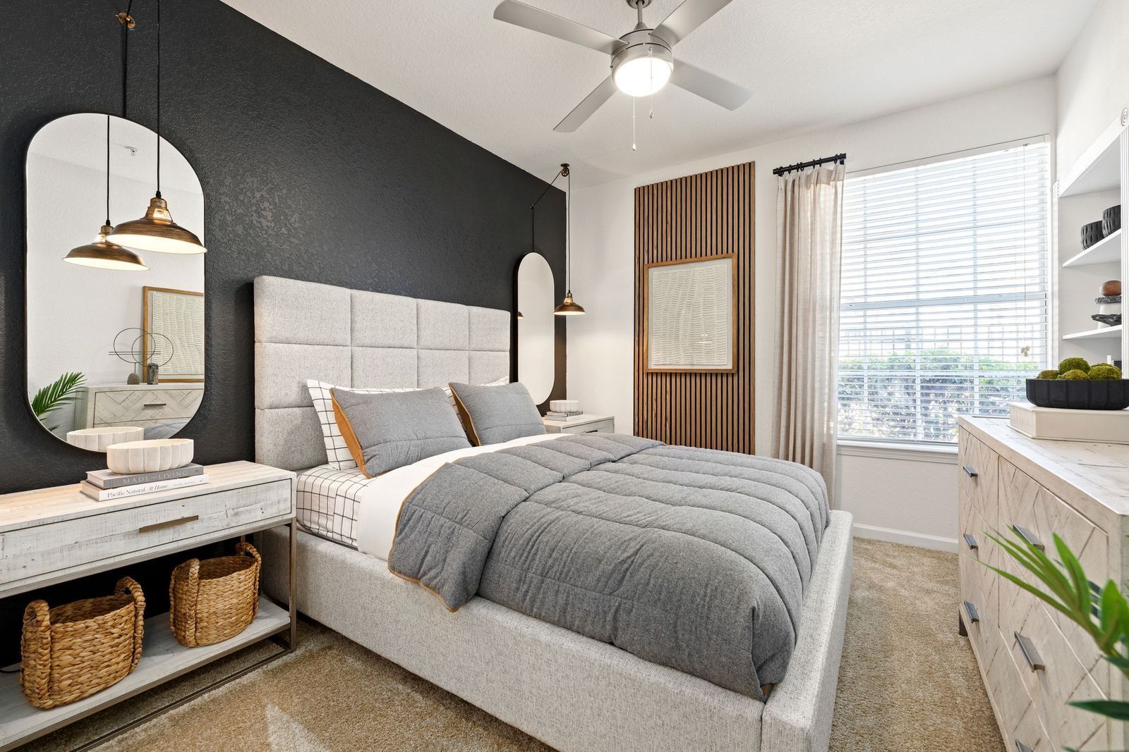 Modern bedroom with a gray tufted bed, black accent wall, oval mirror, white furniture, and a ceiling fan.