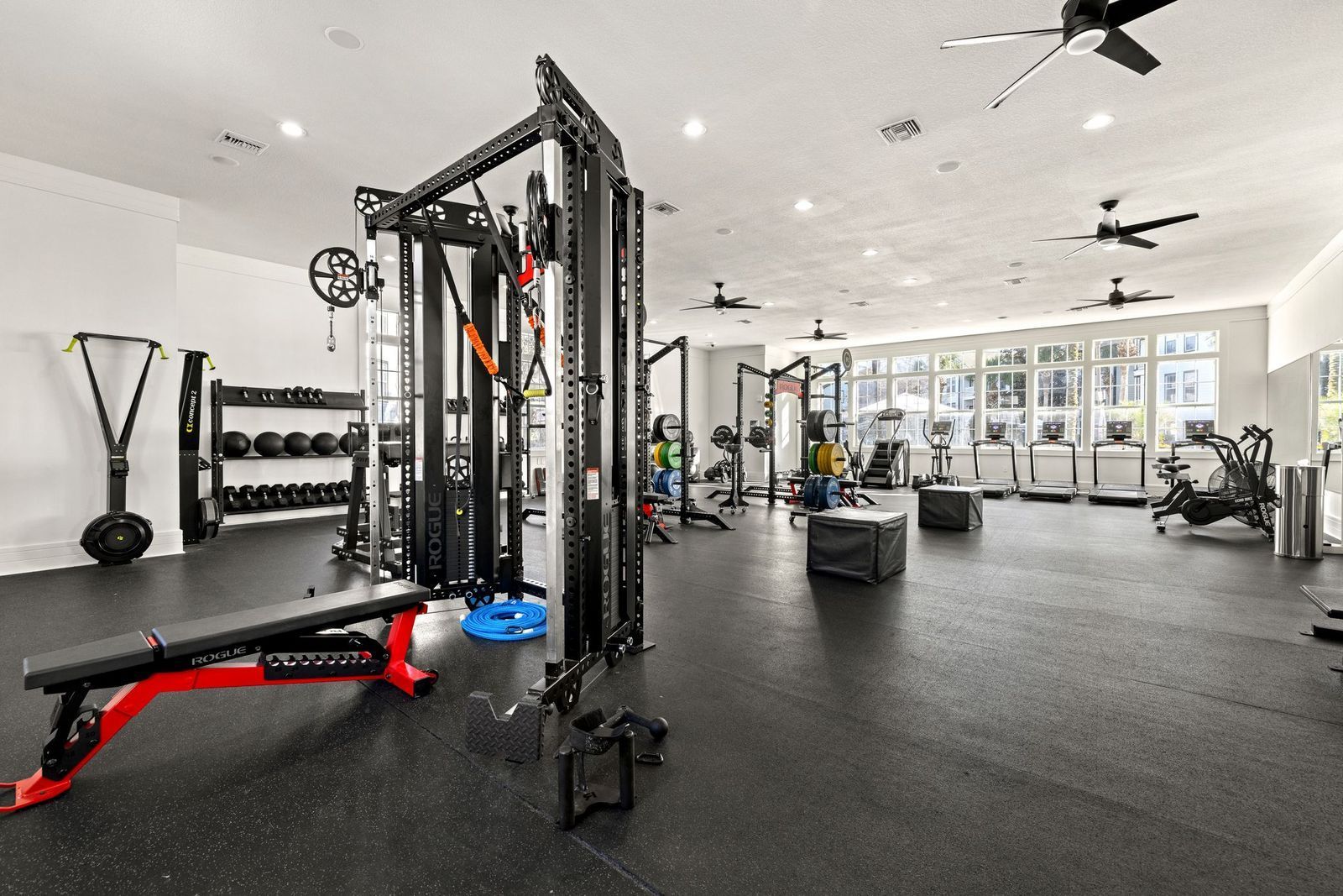 A well-lit, modern gym featuring a black functional trainer, weight racks, treadmills, and a red exercise bench.