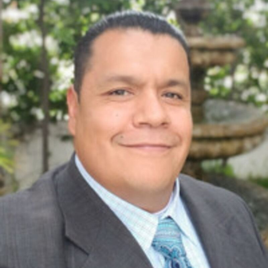 Man in suit smiles outdoors with a water fountain in the background.