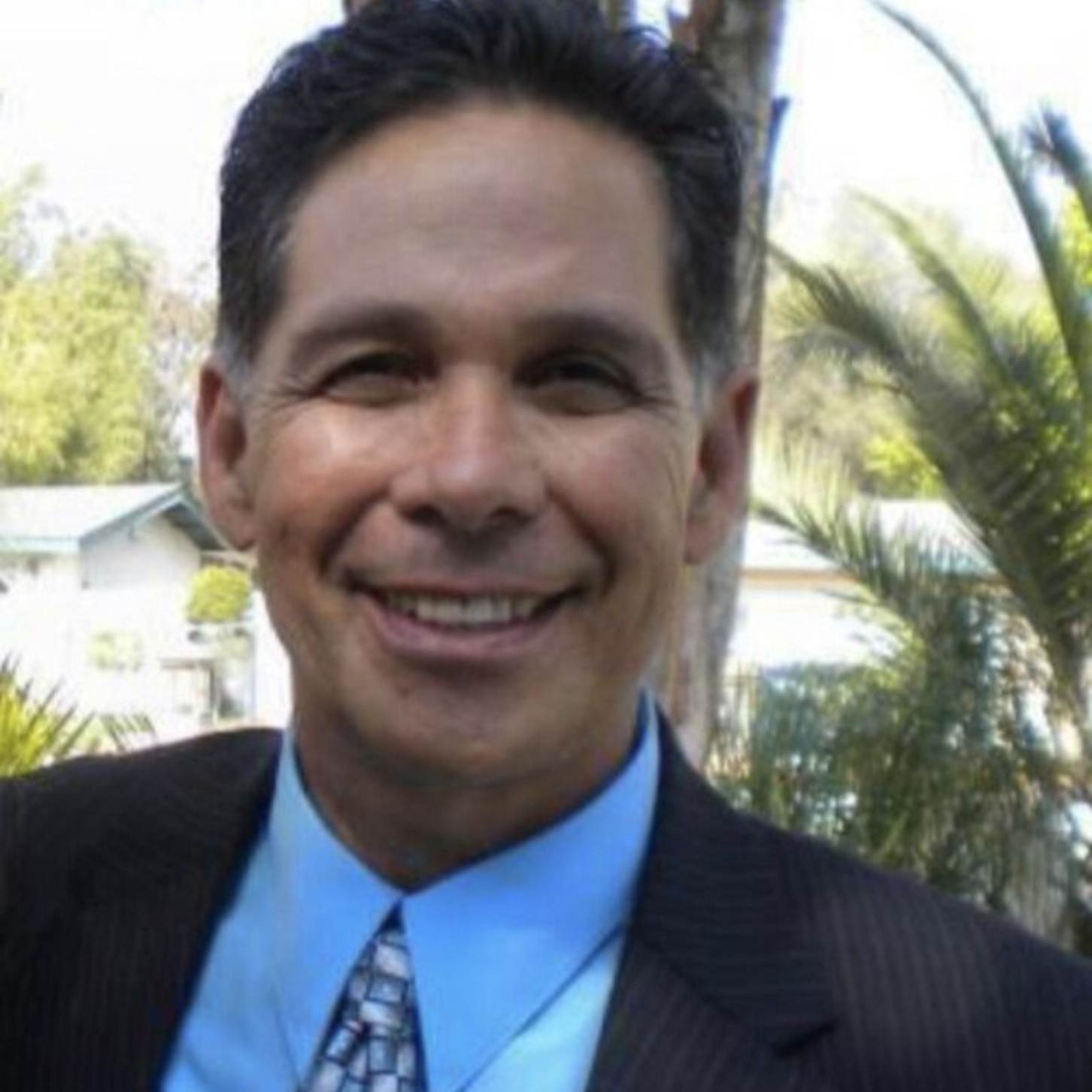 Man in a suit smiles, outdoors. Blue shirt, patterned tie, dark hair.