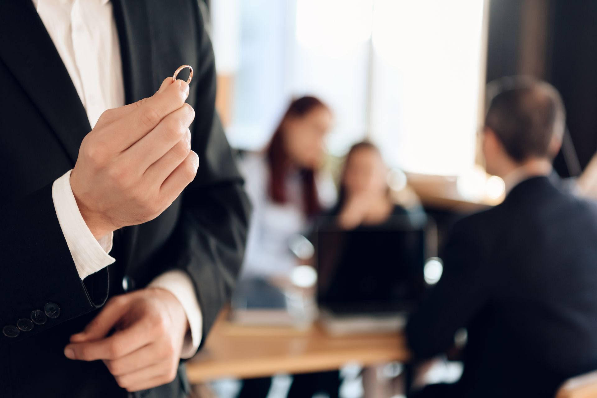 A close-up of a man’s hands holding a ring. A woman, girl, and lawyer are blurred in the background.