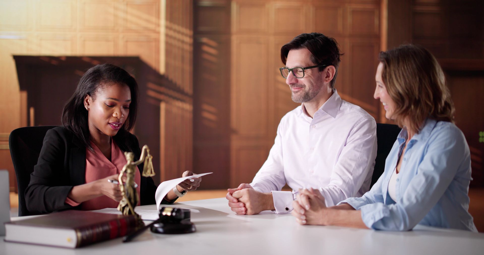 A legal professional reviews documents with two clients seated at a desk in an office setting A legal professional reviews documents with two clients seated at a desk in an office setting