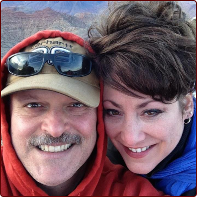 A smiling couple poses for a close-up selfie outdoors, with the man wearing a Carhartt cap and sunglasses, and red hoodie.