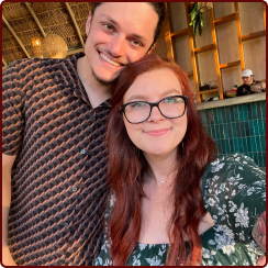 A man with a goatee and a woman with red hair and glasses smile close together in an indoor setting with tiled walls.
