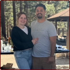 A smiling couple standing together outdoors in a park setting, with trees and a pavilion in the background.