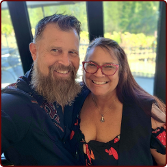 A man with a full beard and a smiling woman wearing glasses pose for a photo in front of a window.