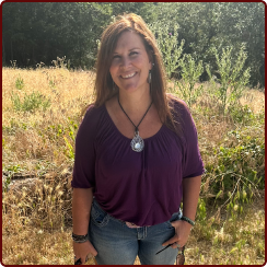 A smiling person in a purple shirt and jeans stands in a field of dry grass with trees in the background.