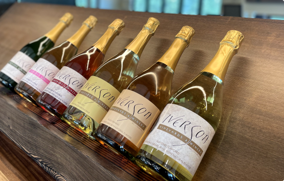 Six bottles of Iverson sparkling wine with various colored labels and liquids arranged in a row on a wooden surface.