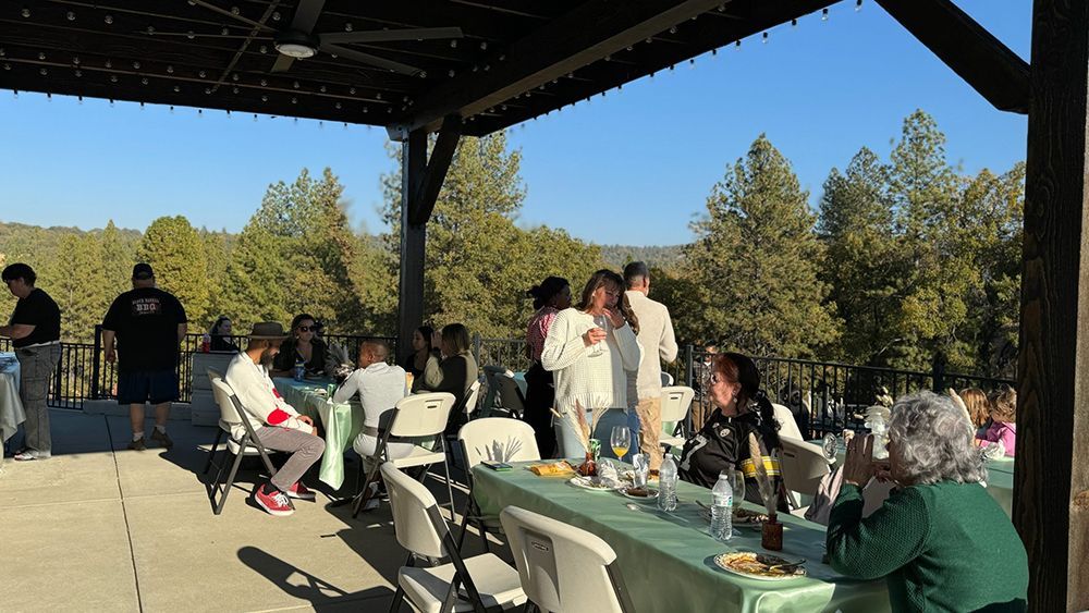 A group of people gather for an outdoor event at tables under a pavilion, with a view of trees in the background.