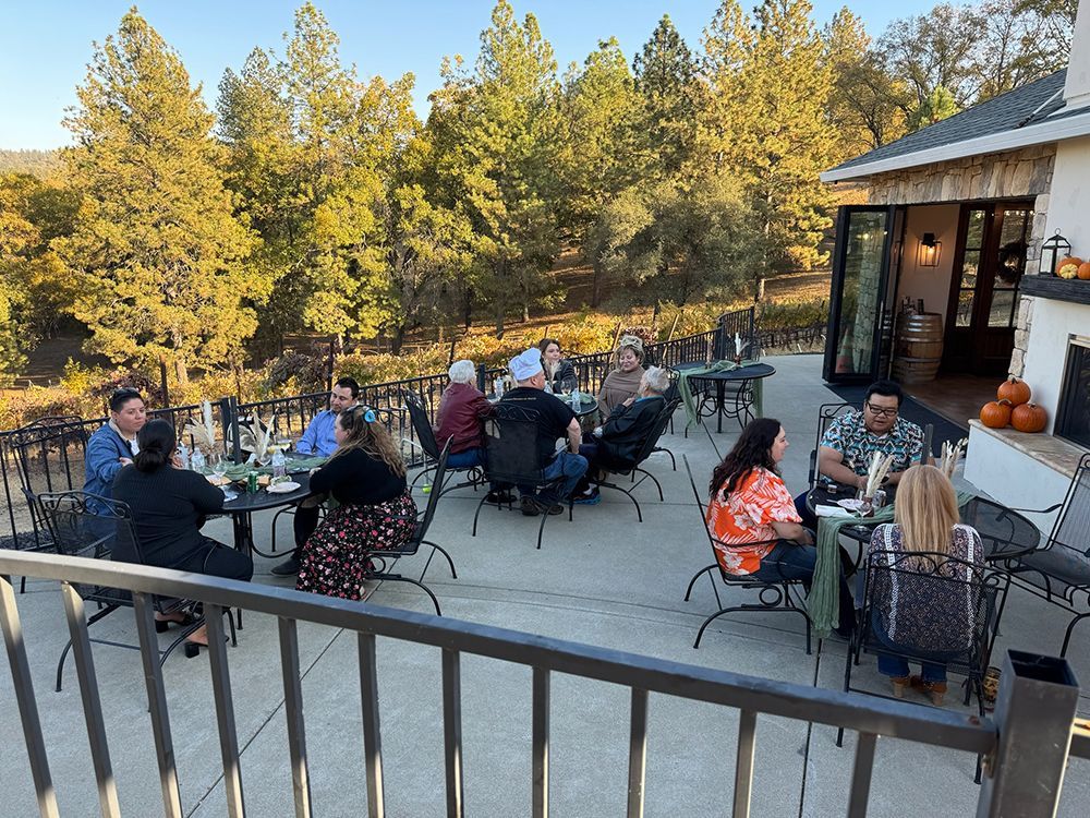 People seated at tables on an outdoor patio at sunset, with a forest in the background and a building to the right.