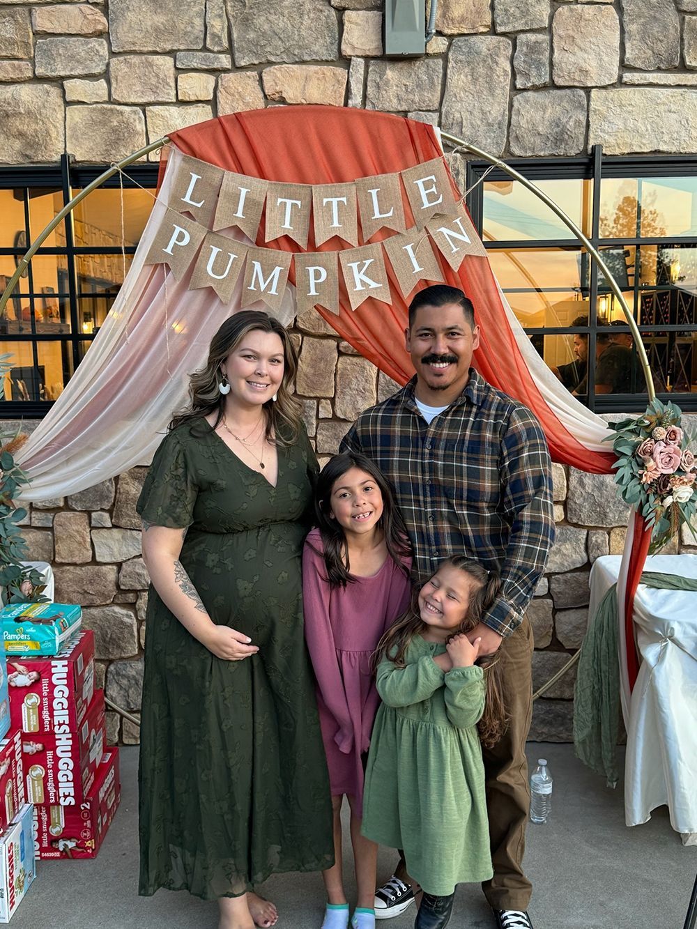 A family stands before a "Little Pumpkin" backdrop for an event, smiling in front of a stone wall.