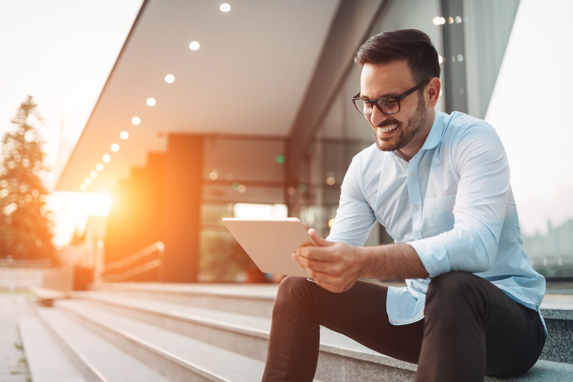 a man wearing glasses is sitting on a set of stairs using a tablet a man wearing glasses is sitting on a set of stairs using a tablet