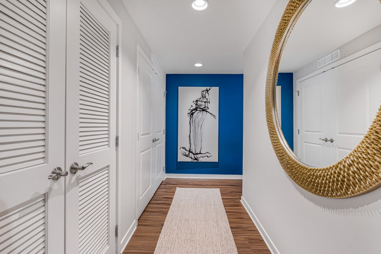 Hallway inside an apartment: white doors, blue accent wall with artwork, and a large woven round mirror.