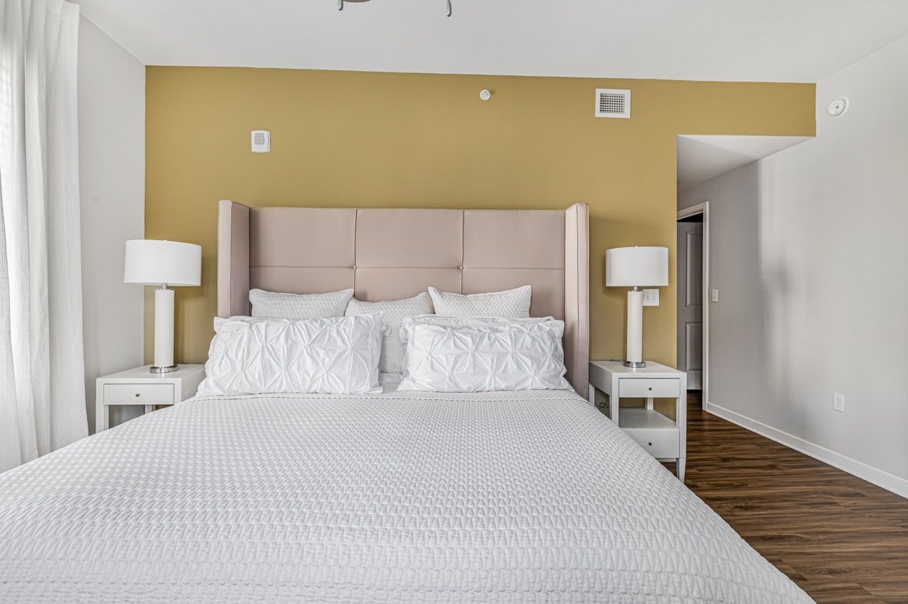Bedroom with an upholstered headboard, white bedding, two nightstands with lamps, and a mustard accent wall.