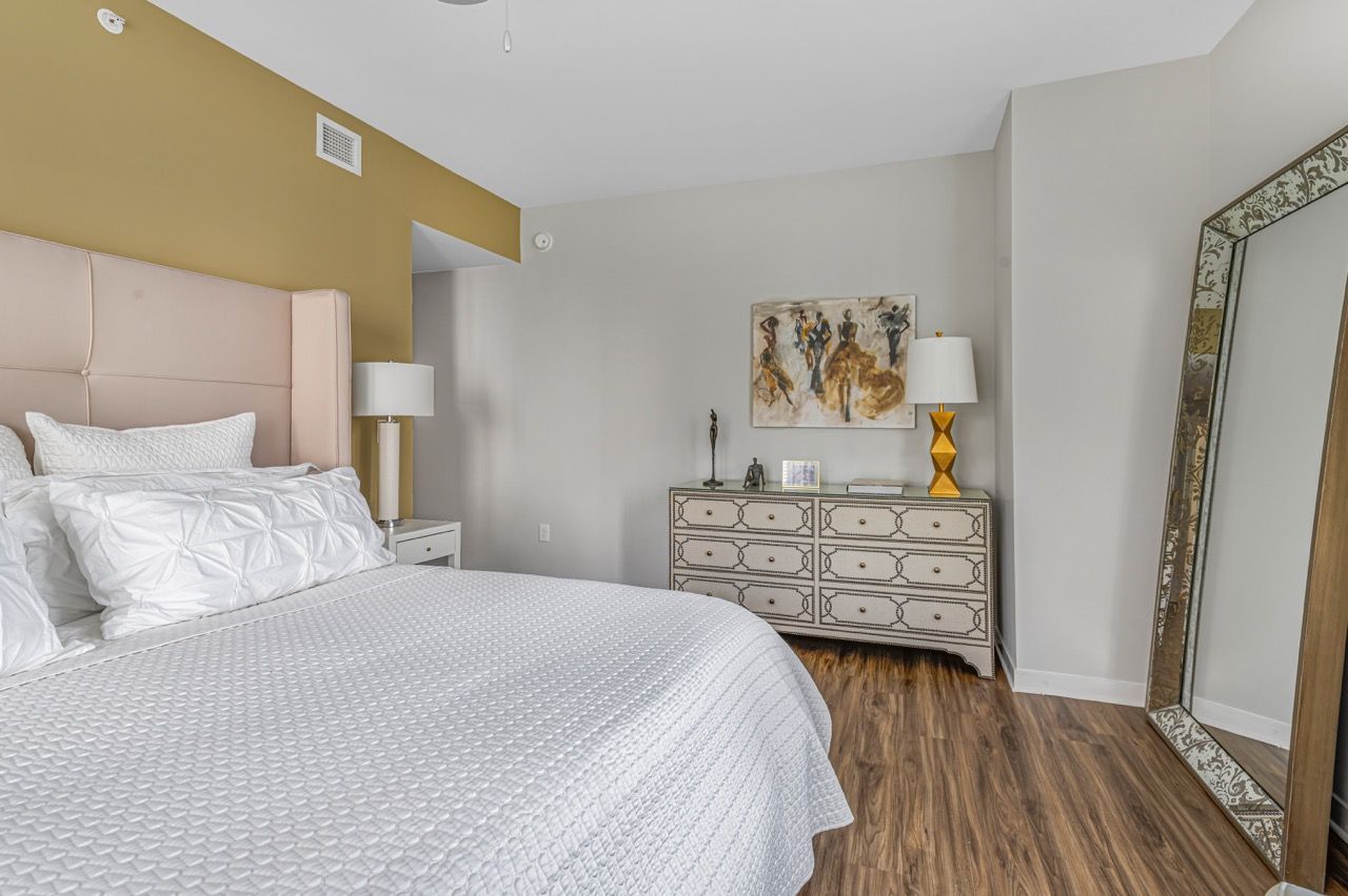 Bedroom in an apartment with a pink upholstered headboard, white bedding, a dresser, and a large ornate full-length mirror.
