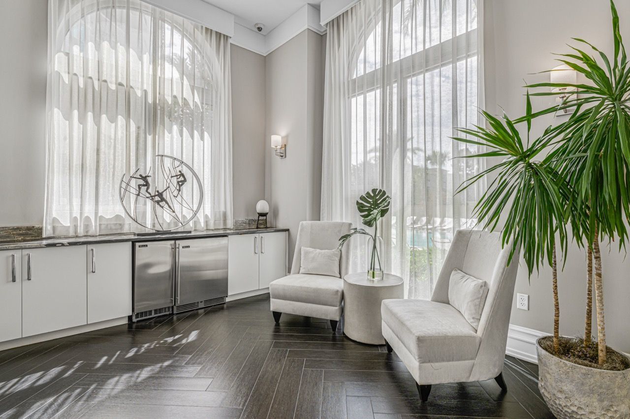 Bright modern living area with white cabinets, stainless steel appliances, sheer curtains, and large potted plant.