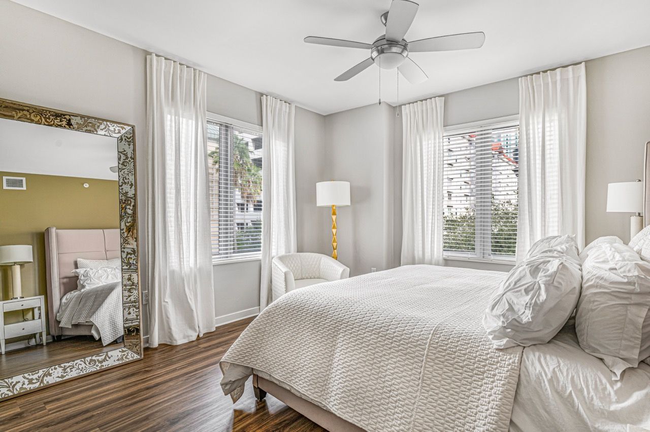 Bedroom with white bedding, mirrored ornate frame, and two windows with white curtains.